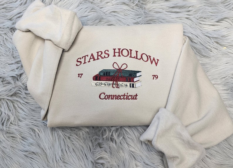 K&ouml;nnte beinhalten: Cremefarbenes Sweatshirt mit gesticktem Schriftzug "STARS HOLLOW" und einem Stapel B&uuml;cher, die mit einer Schleife zusammengebunden sind. Das Design enth&auml;lt die Zahlen "17" und "79" und das Wort "Connecticut".