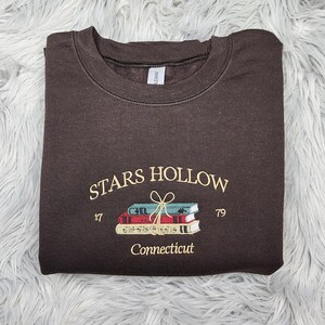 K&ouml;nnte beinhalten: Ein dunkelbraunes Sweatshirt mit gesticktem Text und einem B&uuml;cherstapel. Der Text lautet "STARS HOLLOW 17 79 Connecticut" in cremefarbenem Garn. Die B&uuml;cher sind in Rot-, Blau- und Cremet&ouml;nen gehalten.