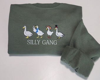 May include: An olive green sweatshirt featuring an embroidered design of four geese, each with unique accessories. The geese are wearing a scarf, a cowboy hat, and a top hat. The text "SILLY GANG" is embroidered below the geese.
