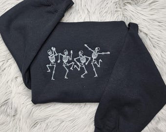 May include: Black sweatshirt featuring a white embroidered design of four dancing skeletons. The skeletons are in various poses, with arms and legs outstretched. The sweatshirt is folded on a white, fluffy surface.