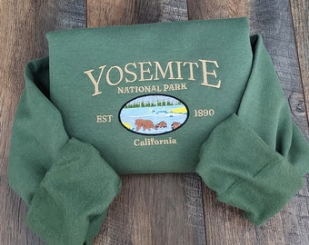 May include: A forest green sweatshirt with the embroidered text "YOSEMITE NATIONAL PARK" and an oval graphic of bears and a lake. The text "EST 1890 California" is also embroidered on the sweatshirt. The sleeves are folded.