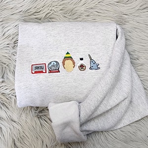 Könnte beinhalten: Ein hellgraues Sweatshirt mit aufgestickten Aufnähern mit Bildern aus dem Film Elf. Zu den Aufnähern gehören das Wort "SANTA!" in Rot, eine Schneekugel, Buddy the Elfs Gesicht, Ahornsirup und ein Narwal.