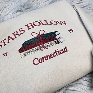 K&ouml;nnte beinhalten: Cremefarbenes Sweatshirt mit burgunderfarbener Stickerei. Das Design zeigt die Worte "STARS HOLLOW Connecticut", einen Stapel B&uuml;cher, die mit einer Schleife zusammengebunden sind, und die Zahlen "17" und "79".