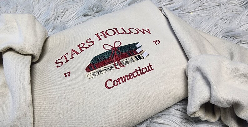K&ouml;nnte beinhalten: Cremefarbenes Sweatshirt mit burgunderfarbener Stickerei. Das Design zeigt die Worte "STARS HOLLOW Connecticut", einen Stapel B&uuml;cher, die mit einer Schleife zusammengebunden sind, und die Zahlen "17" und "79".