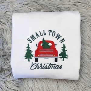 Puede incluir: Sudadera blanca con un dise&ntilde;o bordado que presenta una camioneta roja que transporta un &aacute;rbol de Navidad. El texto "SMALL TOWN CHRISTMAS" est&aacute; arqueado sobre la camioneta, con pinos verdes a cada lado.