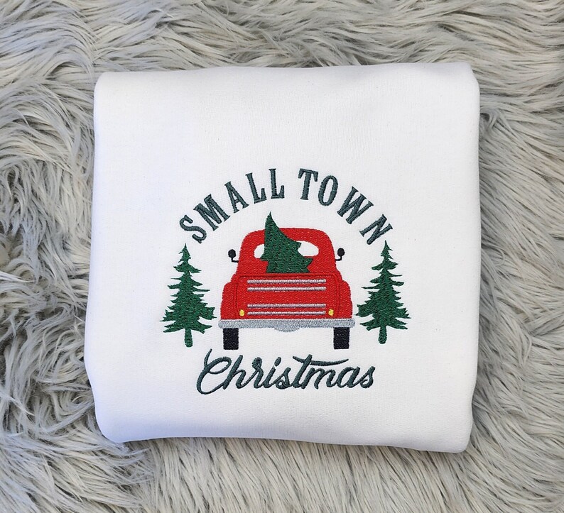Puede incluir: Sudadera blanca con un dise&ntilde;o bordado que presenta una camioneta roja que transporta un &aacute;rbol de Navidad. El texto "SMALL TOWN CHRISTMAS" est&aacute; arqueado sobre la camioneta, con pinos verdes a cada lado.