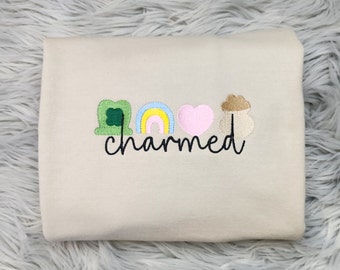 May include: A beige sweatshirt with embroidered St. Patrick's Day themed designs. The design includes a green shamrock, a rainbow, a pink heart, and a pot of gold. The word "charmed" is written in black script below the designs.