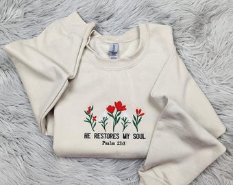 May include: Cream-colored sweatshirt with embroidered red and white flowers and green stems. The text "HE RESTORES MY SOUL" and "Psalm 23:3" are embroidered below the flowers. The sweatshirt has long sleeves.