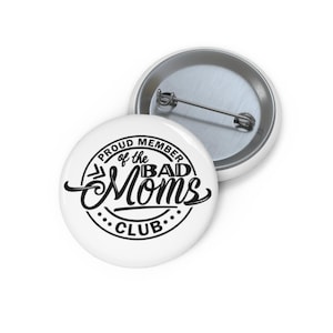 May include: Two white round buttons. One button has the text "PROUD MEMBER of the BAD MOMS CLUB" in black script and a dotted circle border. The other button is silver and has a pin on the back.