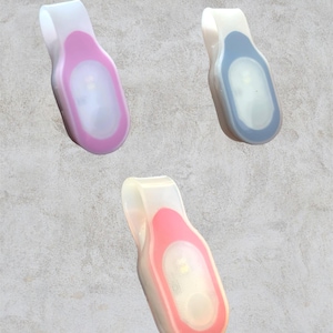May include: Three LED safety lights in pink, blue, and coral. Each light has a white clip and a clear, oval-shaped light. The lights are designed to clip onto clothing or accessories for increased visibility in low-light conditions.