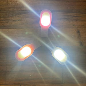 May include: Three small, white LED lights with a curved, rectangular design are arranged on a wooden surface. Each light emits a bright, white glow, with a gradient of color from white to orange or red.