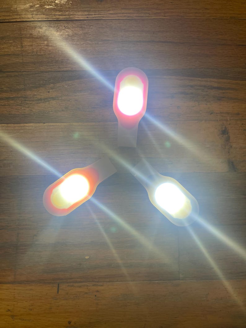 May include: Three small, white LED lights with a curved, rectangular design are arranged on a wooden surface. Each light emits a bright, white glow, with a gradient of color from white to orange or red.