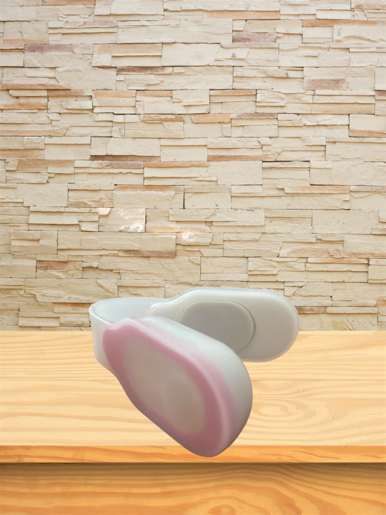 May include: Two white and pink massage tools are displayed on a wooden surface. The tools have a curved design with a rounded end. The background features a light brown brick wall.