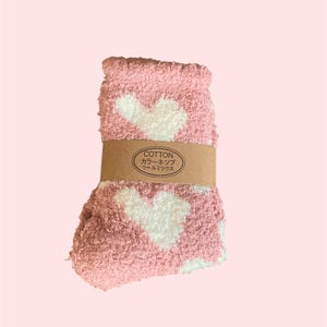 May include: A pair of fuzzy pink socks featuring a white heart pattern. The socks are folded and secured with a brown paper band that has the text COTTON, followed by Japanese characters, printed in black on the front of the paper label.