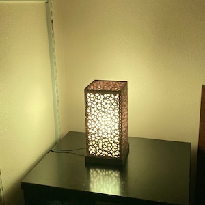 May include: A wooden table lamp with a geometric patterned shade. The lamp is switched on and emits a warm white light.