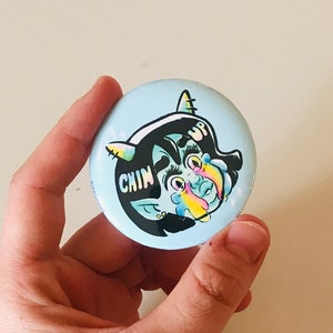 Op de afbeelding: Een blauwe en witte knop met een cartoonillustratie van een blauw wezen met een enkele hoorn, dat een wit hoofdband draagt met het woord "UP" erop. Het wezen heeft een roze neus en blauwe ogen. De tekst "CHIN" staat op de knop.