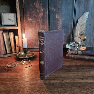 Op de afbeelding: Een donkerpaars hardcover boek getiteld Tales of Mystery and Imagination van Edgar Allan Poe met gouden letters op de rug. Het boek staat rechtop op een houten oppervlak naast een brandende witte kaars, een metalen bril en een ganzenveer.