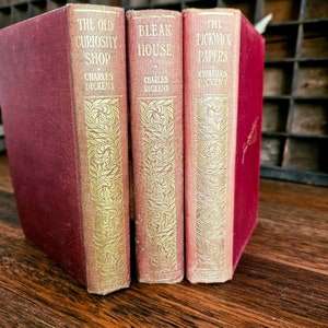Puede incluir: Tres libros antiguos de tapa dura de Charles Dickens. Los libros son rojos con letras doradas y dise&ntilde;os decorativos. Los t&iacute;tulos son "The Old Curiosity Shop", "Bleak House" y "The Pickwick Papers".