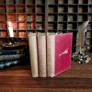 Puede incluir: Tres libros antiguos de tapa dura de Charles Dickens: "The Old Curiosity Shop", "The Bleak House" y "The Pickwick Papers". Los libros est&aacute;n dispuestos en una fila sobre una superficie de madera. Un libro rojo con la palabra "Gadshill" en la portada est&aacute; a la derecha de los libros.