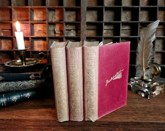 May include: Three vintage hardcover books by Charles Dickens: "The Old Curiosity Shop", "The Bleak House", and "The Pickwick Papers". The books are arranged in a row on a wooden surface. A red book with the word "Gadshill" on the cover is to the right of the books.