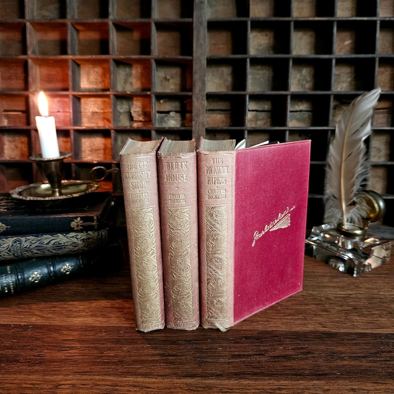 Puede incluir: Tres libros antiguos de tapa dura de Charles Dickens: "The Old Curiosity Shop", "The Bleak House" y "The Pickwick Papers". Los libros est&aacute;n dispuestos en una fila sobre una superficie de madera. Un libro rojo con la palabra "Gadshill" en la portada est&aacute; a la derecha de los libros.