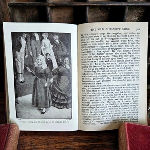 Puede incluir: Una imagen en blanco y negro de un libro antiguo que representa una escena de la novela "The Old Curiosity Shop" de Charles Dickens. La imagen muestra a un grupo de personas de pie en un escenario, incluida una ni&ntilde;a que sostiene un palo. El texto "Mrs. Jarley was at great pains to instruct her." es visible en la parte inferior de la imagen.
