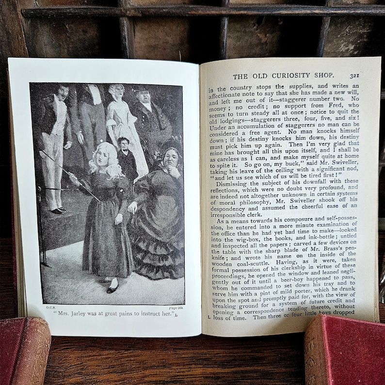 Puede incluir: Una imagen en blanco y negro de un libro antiguo que representa una escena de la novela "The Old Curiosity Shop" de Charles Dickens. La imagen muestra a un grupo de personas de pie en un escenario, incluida una ni&ntilde;a que sostiene un palo. El texto "Mrs. Jarley was at great pains to instruct her." es visible en la parte inferior de la imagen.