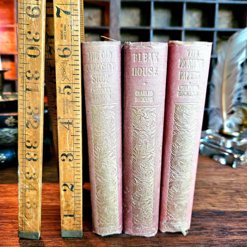 Puede incluir: Tres libros antiguos de tapa dura con cubiertas rojas y letras doradas. Los t&iacute;tulos son "The Old Curiosity Shop", "Bleak House" y "The Pickwick Papers". Los tres libros son de Charles Dickens.