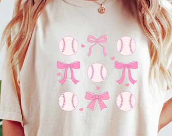 May include: An ivory t-shirt featuring a repeating grid pattern of pink baseballs and pink bows. The graphic design includes small pink hearts interspersed between the baseballs and bows, creating a symmetrical arrangement on the front of the shirt.