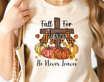 Puede incluir: Una camiseta color crema suave con un gráfico de una cruz de madera drapeada con tela a cuadros, rodeada de calabazas y hojas de otoño. El texto dice Fall For Jesus He Never Leaves. La prenda se combina con jeans azules y un bolso de color beige.