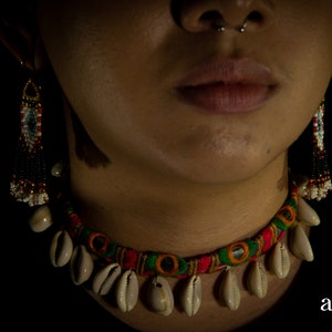 Può includere: Primo piano di una persona che indossa una collana girocollo colorata con conchiglie e orecchini di perline. La collana presenta tessuto rosso, verde e giallo con conchiglie attaccate. Gli orecchini hanno un design di perline multicolore. Il marchio "antribe jewellery" è visibile.