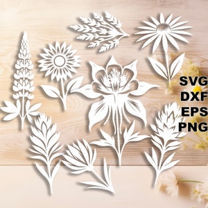 May include: A set of eight white paper cut flower designs, including a sunflower, a lupine, a columbine, a daisy, and a few other floral designs. The designs are cut from white paper and are arranged on a wooden background.