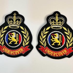 May include: Two embroidered patches with a black background, gold crown, and laurel leaves. A central emblem features a yellow lion, red and blue accents, and the word "UNDERCOVER" in red. These patches are likely for uniforms or collectibles.