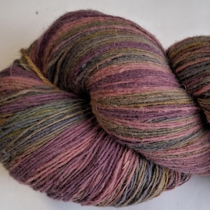 Puede incluir: Un ovillo de lana en tonos morado, rosa, verde y gris. La lana está bien enrollada, mostrando la textura de las fibras. Los colores se mezclan, creando un efecto jaspeado, adecuado para proyectos de tejido o ganchillo.