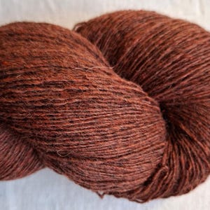 May include: A skein of yarn in a deep, rich shade of reddish-brown. The fiber has a visible, slightly fuzzy texture with subtle variations in tone throughout the twisted strands, creating a consistent, earthy appearance across the entire length.