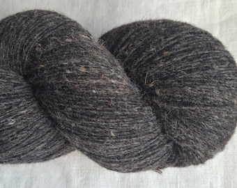 May include: A skein of dark charcoal gray yarn with a textured, slightly fuzzy appearance. The yarn features subtle flecks of lighter gray and brown fibers throughout the twisted strands, creating a heathered effect across the entire surface.