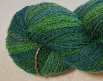 May include: A skein of yarn in shades of green and teal. The yarn is tightly wound and secured with a brown twine. The colors blend together, creating a textured appearance. Ideal for knitting or crocheting projects.