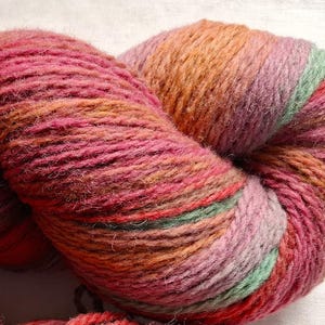 Puede incluir: Primer plano de una madeja de lana colorida. La lana es una mezcla de rosas, naranjas y verdes, creando un efecto degradado. La lana está bien enrollada y parece suave y texturizada, ideal para proyectos de tejido o ganchillo.