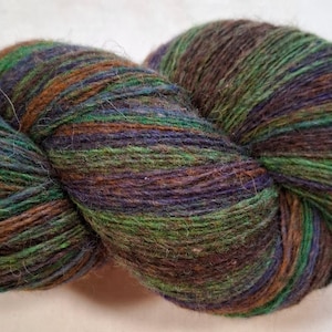 Puede incluir: Un ovillo de lana en una mezcla de colores que incluye verde, marrón, morado y azul. La lana está bien enrollada, mostrando la textura y las variaciones de color. Ideal para proyectos de tejido o ganchillo.