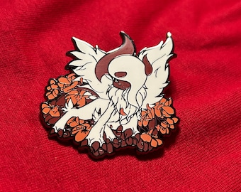 Puede incluir: Un pin de esmalte blanco con una criatura de estilo de dibujos animados con pelaje marrón y una cara blanca. La criatura está rodeada de flores naranjas. El pin está sobre un fondo de tela roja.