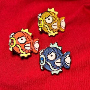 May include: Three enamel pins shaped like a cartoon fish with a yellow, orange, and blue glitter finish. Each fish has a large, round eye and a small mouth with a single tooth. The fish are wearing a yellow, orange, and blue snorkel, respectively.