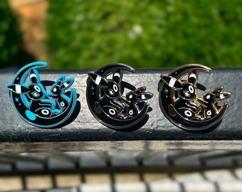 Puede incluir: Tres pines de esmalte con una silueta en blanco y negro de dos evoluciones de Eevee, Espeon y Umbreon, acurrucados en una luna creciente. Los pines están sobre una superficie negra.