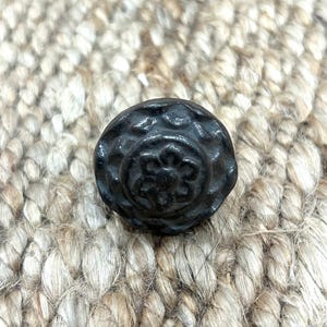 Op de afbeelding: Een donkere, ronde metalen knop met een bloemenontwerp. De knop heeft een verhoogd, getextureerd oppervlak en is geplaatst tegen een geweven, beige achtergrond. De diameter van de knop is ongeveer 3,8 cm.