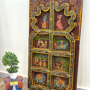 May include: A pair of intricately carved wooden doors featuring colourful painted scenes of people and elephants. The doors are decorated with floral patterns and have a dark brown finish.