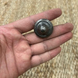 May include: A small, round, metal knob with a domed top and a raised ring around the edge. The knob is a dark grey colour.