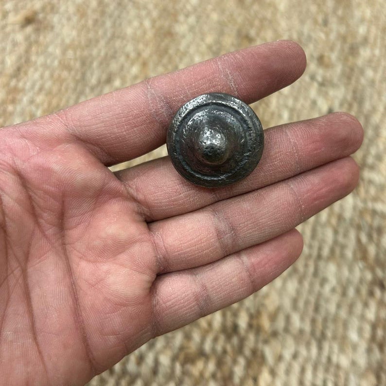 May include: A small, round, metal knob with a domed top and a raised ring around the edge. The knob is a dark grey colour.