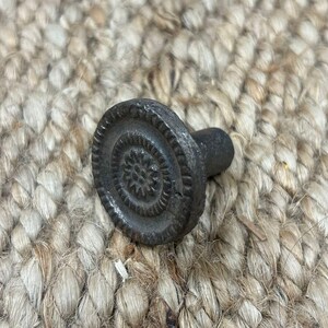 Op de afbeelding: Een zwarte metalen knop met een cirkelvormig ontwerp en een verhoogd midden. De knop heeft een getextureerd oppervlak en een kleine, ronde steel.