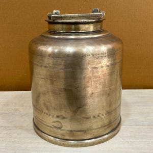 Puede incluir: Un recipiente de latón vintage con una tapa con bisagras y un diseño decorativo. El recipiente tiene una forma cilíndrica y un acabado liso y envejecido.