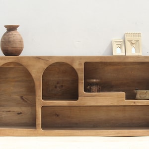 Puede incluir: Una estantería de madera con aberturas arqueadas y varios estantes. La estantería está hecha de madera marrón claro y tiene un acabado natural. Hay pequeños objetos decorativos en los estantes.
