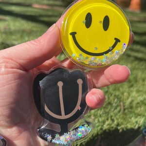 May include: Two clear plastic round ornaments with glitter inside. One ornament has a yellow smiley face and the other has a black silhouette of a flower.